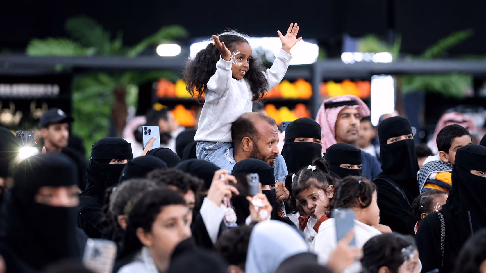 Crowd gathered at an outdoor event, with a child sitting on an adult’s shoulders and families capturing the moment on smartphones.