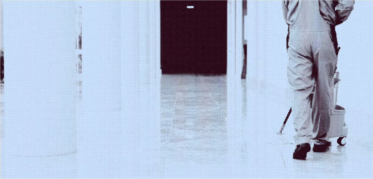 Person wearing work clothes pushing a mop bucket in a hallway with a tiled floor.