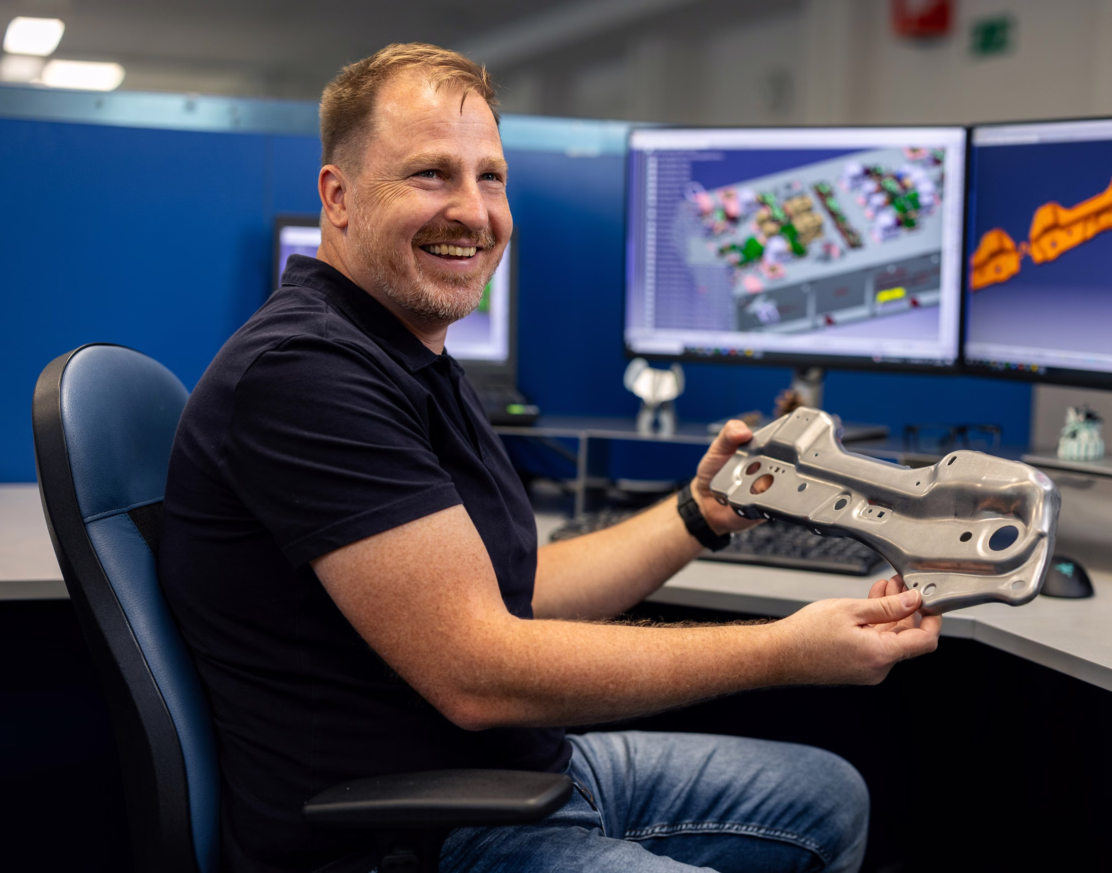 Design engineer holding a formed sheet-metal part beside CAD workstations