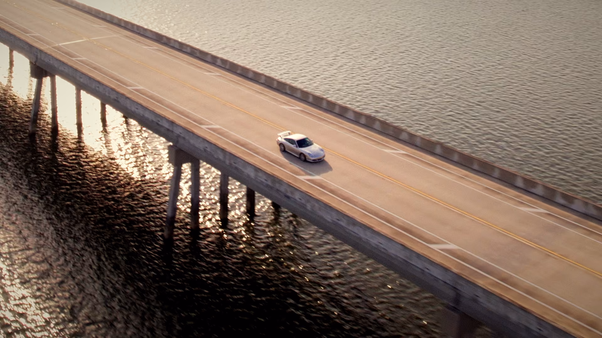 Silver sports car driving alone on a bridge over calm water during sunset.