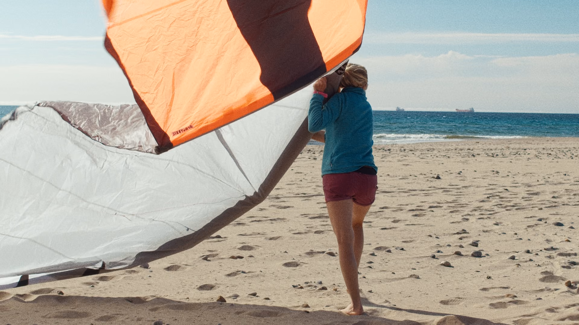 Person holding a large orange and white kite on a sandy beach near the ocean.