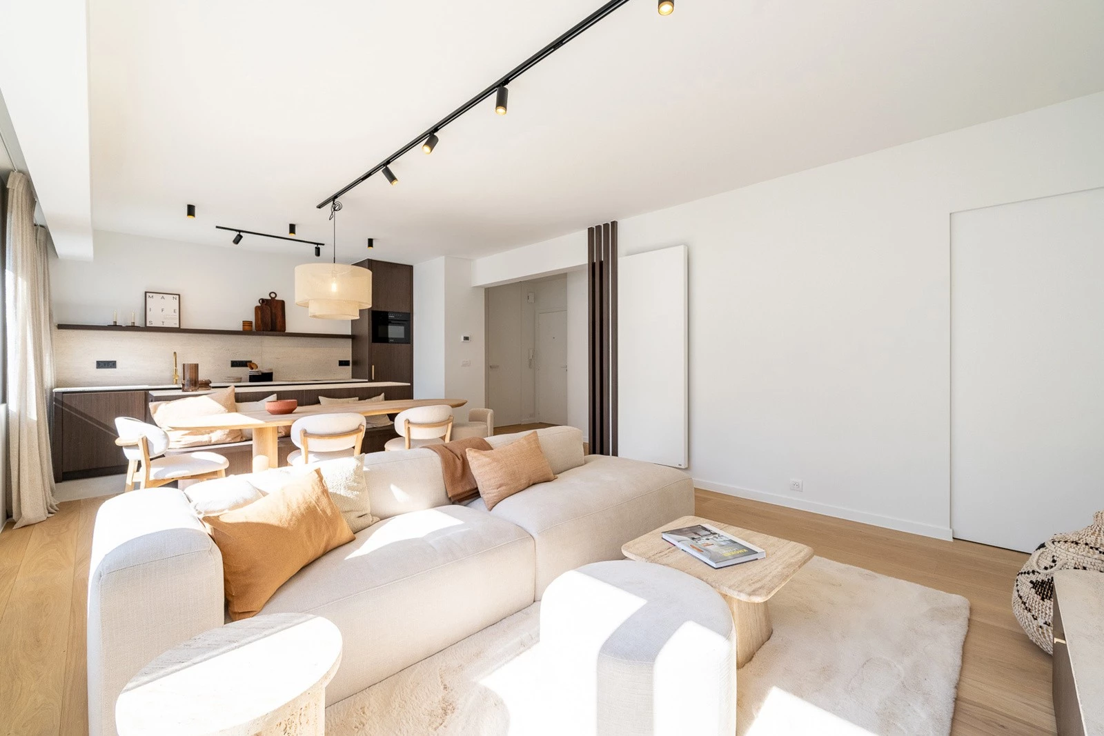 Bright modern open living and dining area with light wood floors, white walls, beige sectional sofa with brown and beige pillows, small wooden coffee table, and an open kitchen with dark wood cabinets, a dining table, and chairs, illuminated by natural light from a window on the left side.
