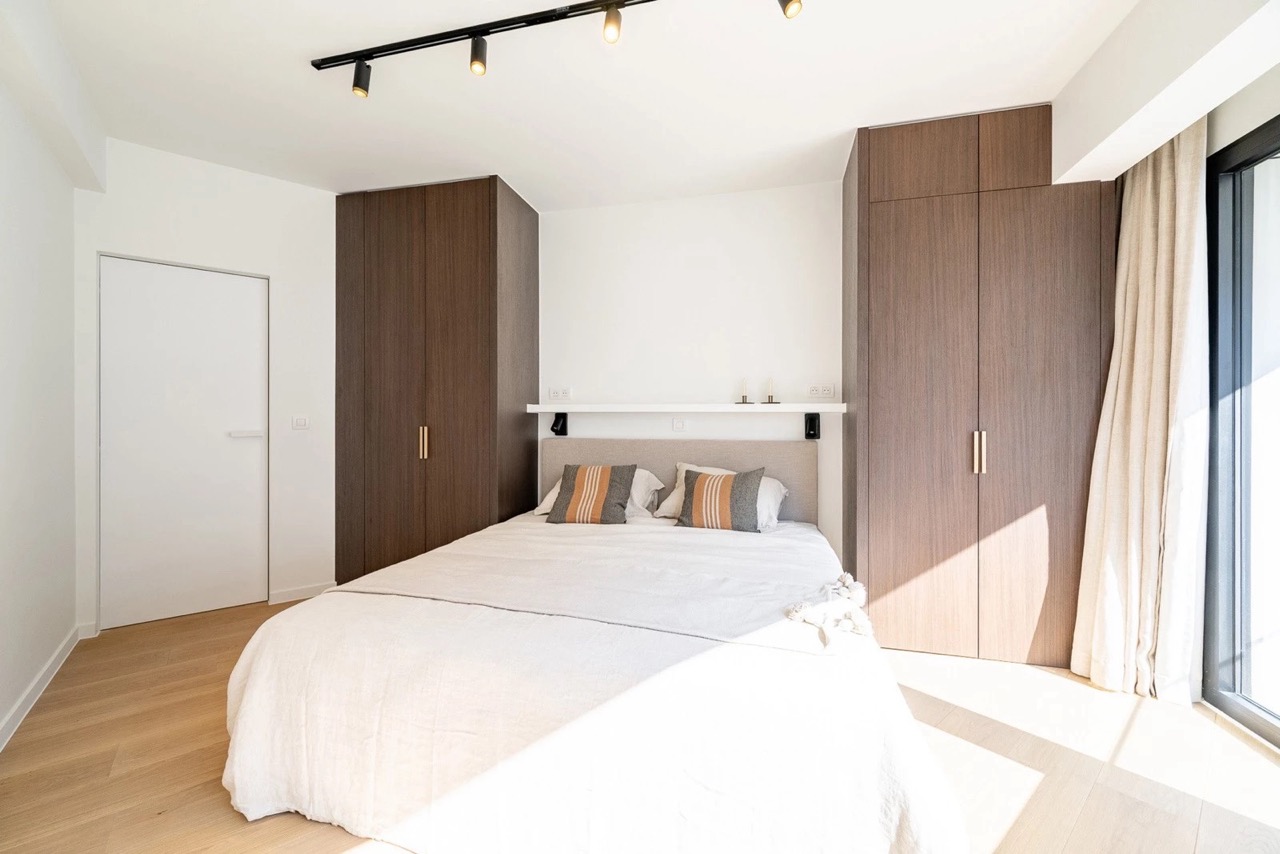 Bright modern bedroom with a large bed covered in white linens and two pillows with gray and orange stripes, flanked by two tall wooden wardrobes, featuring a white shelf above the bed with minimal decor and large window with beige curtains letting in natural light.