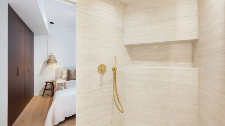 Minimalist shower area with beige stone walls, a built-in shelf, and a gold-tone showerhead and control; a view into a bedroom with a bed, wooden nightstand, large dark wardrobe, and woven hanging lamp.