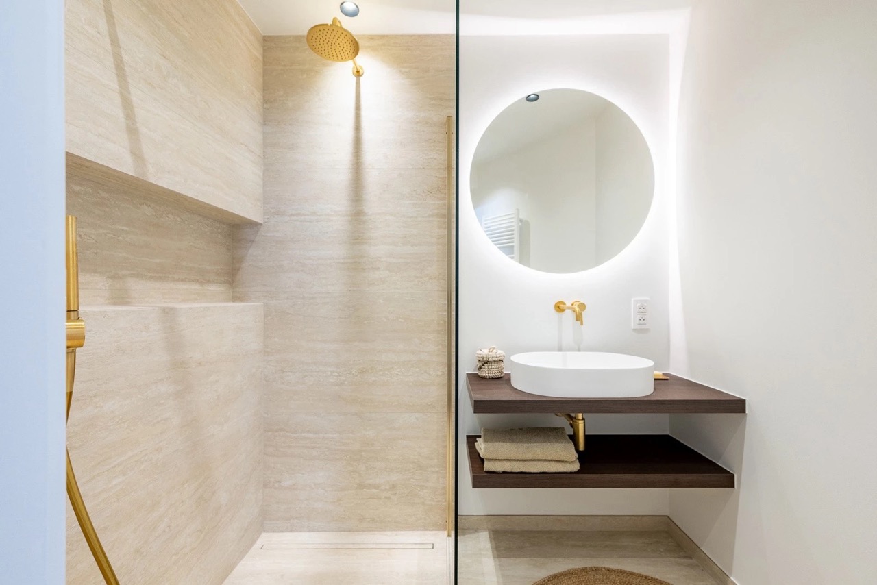 Modern bathroom with beige tiled walk-in shower featuring a gold rain showerhead and a separate handheld shower. Opposite the shower is a floating dark wood vanity with a white round vessel sink, gold faucet, and a round backlit mirror above, with towels stored on the lower shelf.
