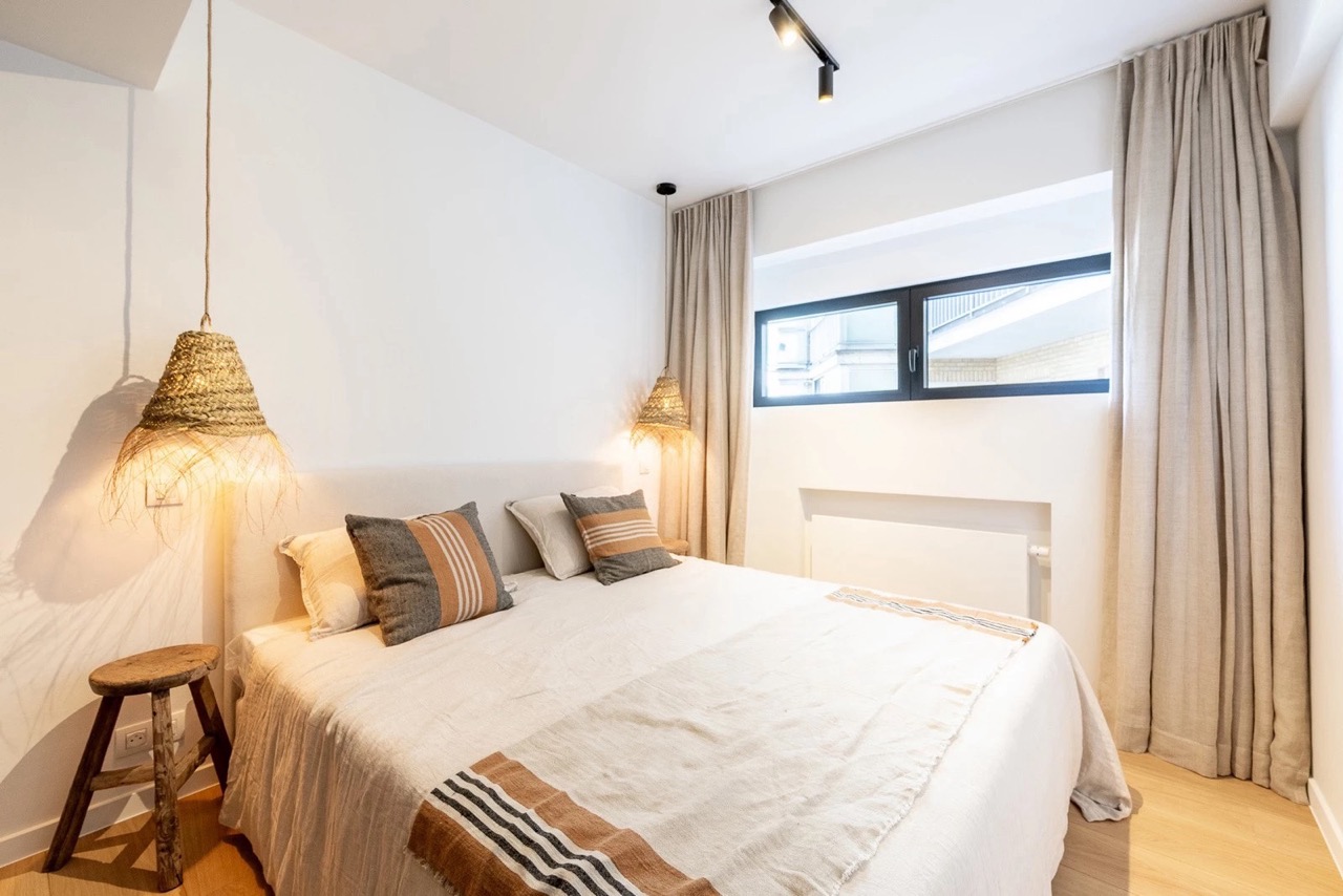 Minimalist bedroom with a large bed covered in light-colored bedding with striped pillows and a matching throw. Two woven pendant lamps hang on either side of the bed, beside a small rustic wooden stool. The room features light wooden flooring, beige curtains, and a narrow horizontal window with a black frame letting in daylight.