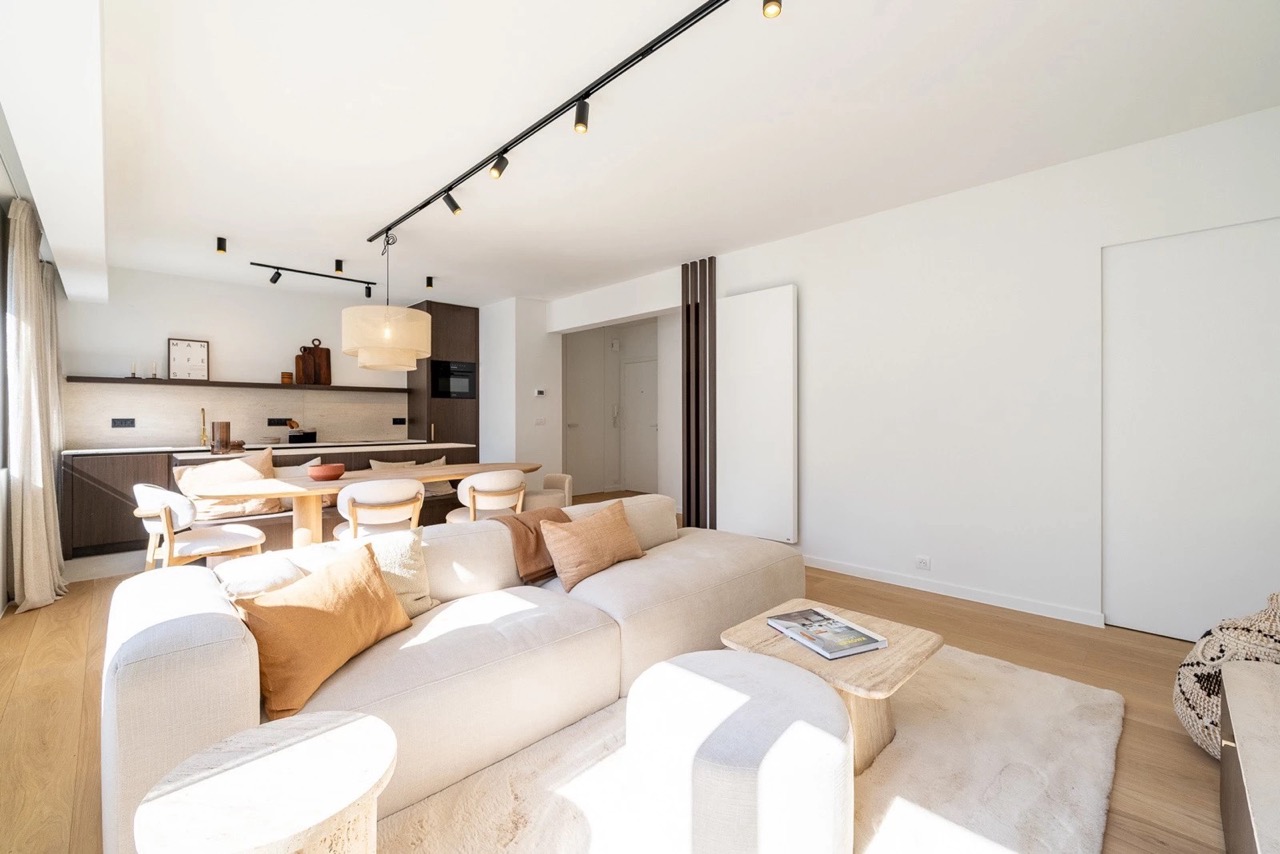 Bright, modern open-plan living space with a beige sofa adorned with tan and cream pillows, a small light wood coffee table with a magazine, a dining area with six chairs around a wooden table, dark wood kitchen cabinets, a white pendant light, and large windows with sheer curtains letting in natural light.