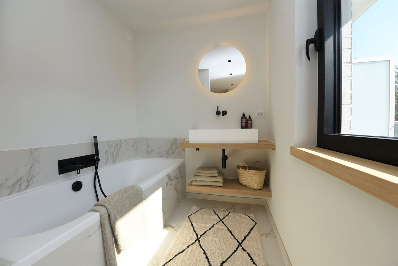 Minimalist bathroom with a white bathtub featuring black fixtures and a beige towel draped over the side, a wall-mounted wooden shelf with a white rectangular sink, hand soap bottles, folded towels, and a woven basket underneath, illuminated round mirror above the sink, and a window with a wooden sill letting in natural light.
