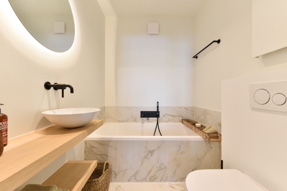 Minimalist bathroom with white and light wood tones featuring a rectangular bathtub with marble tile surround, black faucet and hand shower, a white vessel sink on a wooden shelf, a round mirror with backlight, and a white toilet with dual flush buttons.