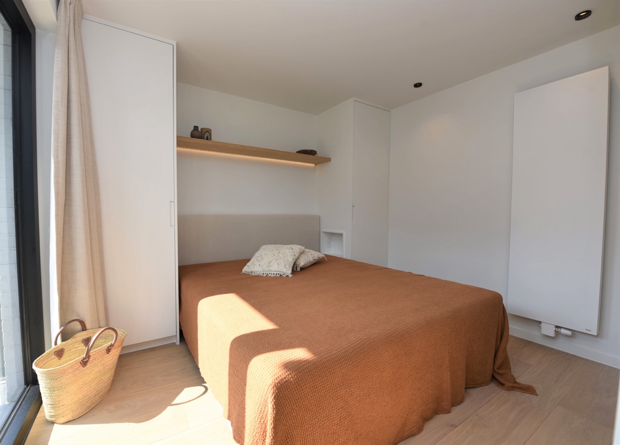 Minimalist bedroom with a large bed covered in a brown blanket and two patterned pillows, flanked by two white wardrobes, a floating wooden shelf with decorative items above the bed, a woven basket on the light wooden floor near a window with beige curtains, and a wall-mounted white radiator on the right.