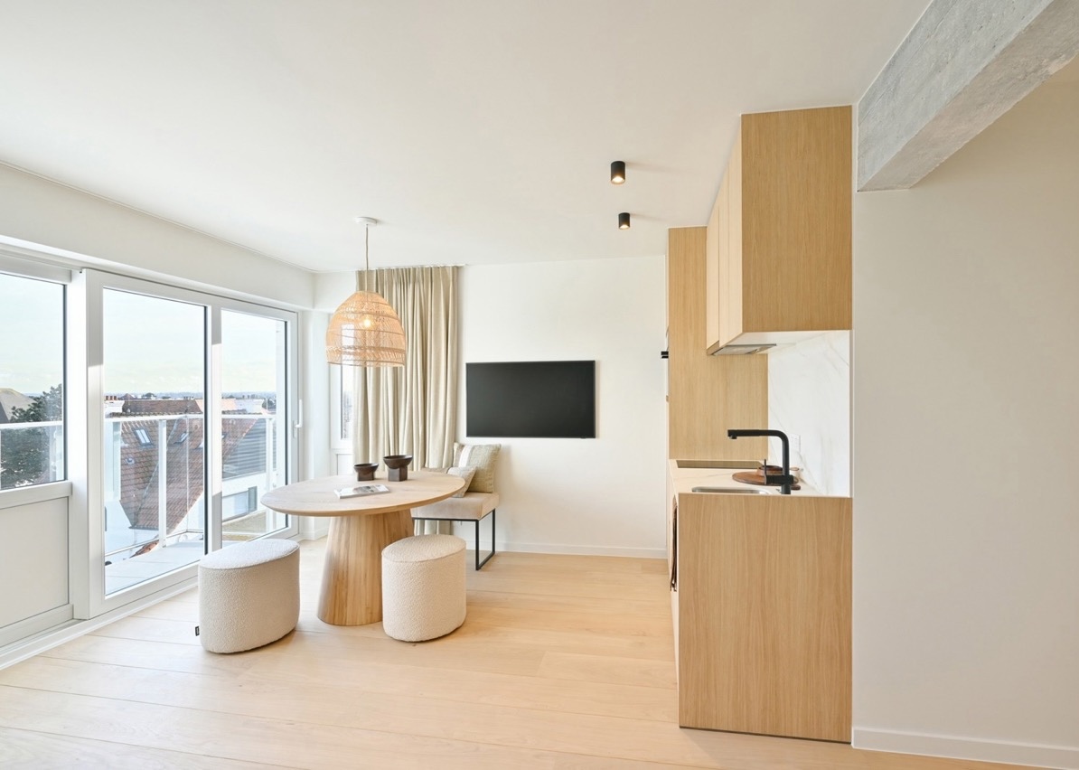 Bright modern kitchen and dining area with light wood floor and cabinets, a round wooden table with beige cushioned stools, a hanging wicker lampshade, large sliding glass doors letting in natural light, a wall-mounted flat screen TV, and a beige curtain covering a corner window.