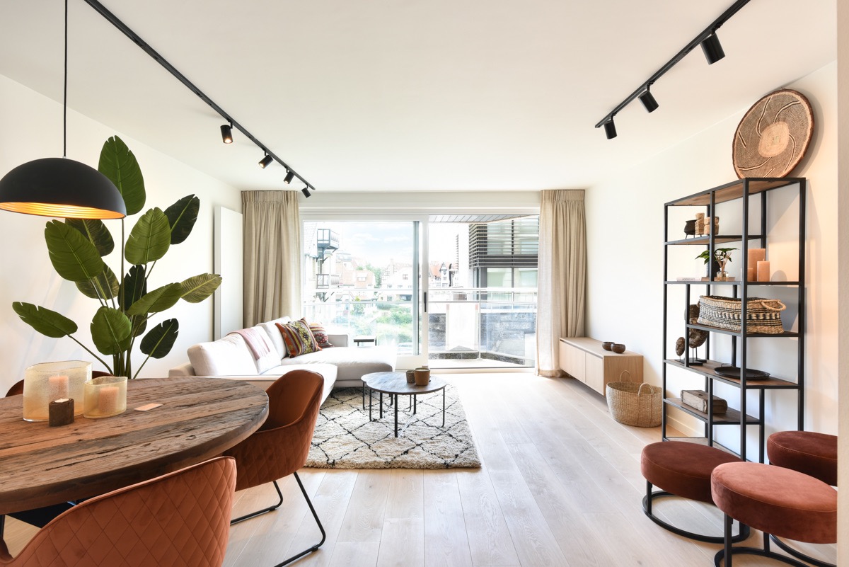 Bright modern living room with large sliding glass doors leading to a balcony, a white sectional sofa with colorful pillows on a patterned rug, a round wooden dining table with burnt orange chairs, tall green plant under a hanging black lamp, and a black metal shelving unit with decorative items on the right.