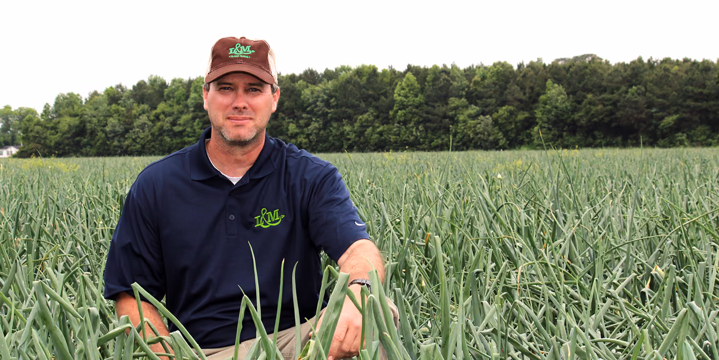 Man wearing a brown cap and navy blue polo shirt with green logo, sitting in a dense green onion field with a background of trees.