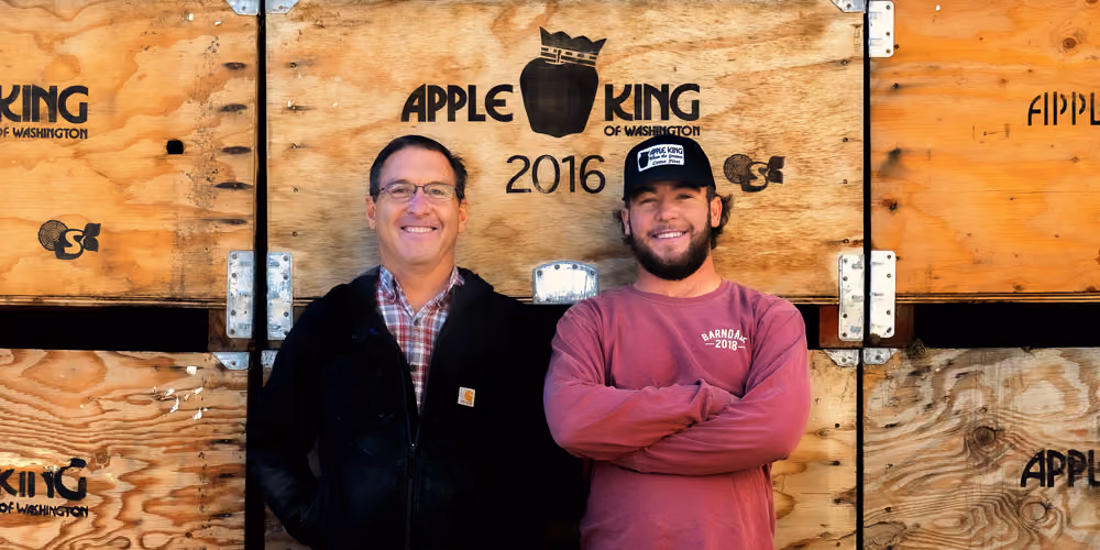 Two men standing and smiling in front of large wooden crates marked 'Apple King of Washington 2016' with an apple wearing a crown logo.