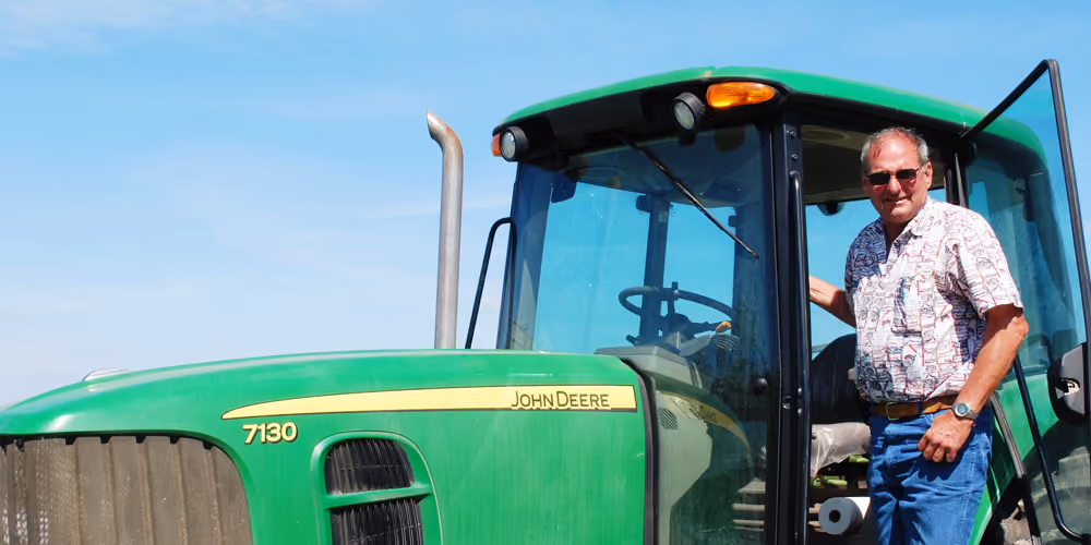 Man wearing sunglasses, a patterned short-sleeve shirt, and blue jeans standing beside a green John Deere 7130 tractor under a clear blue sky.