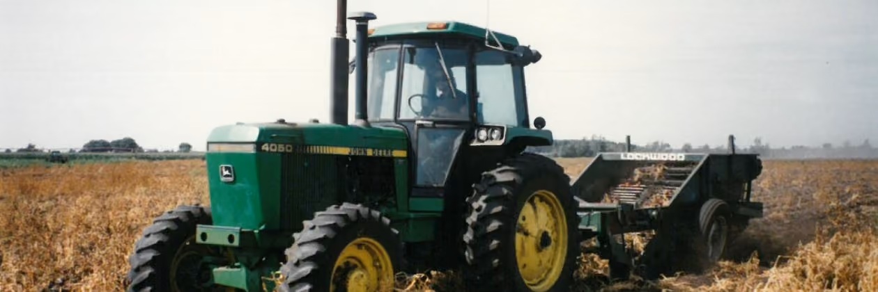 Green John Deere 4050 tractor with large yellow wheels pulling a Rootwood trailer across a field of dry crops under a clear sky.