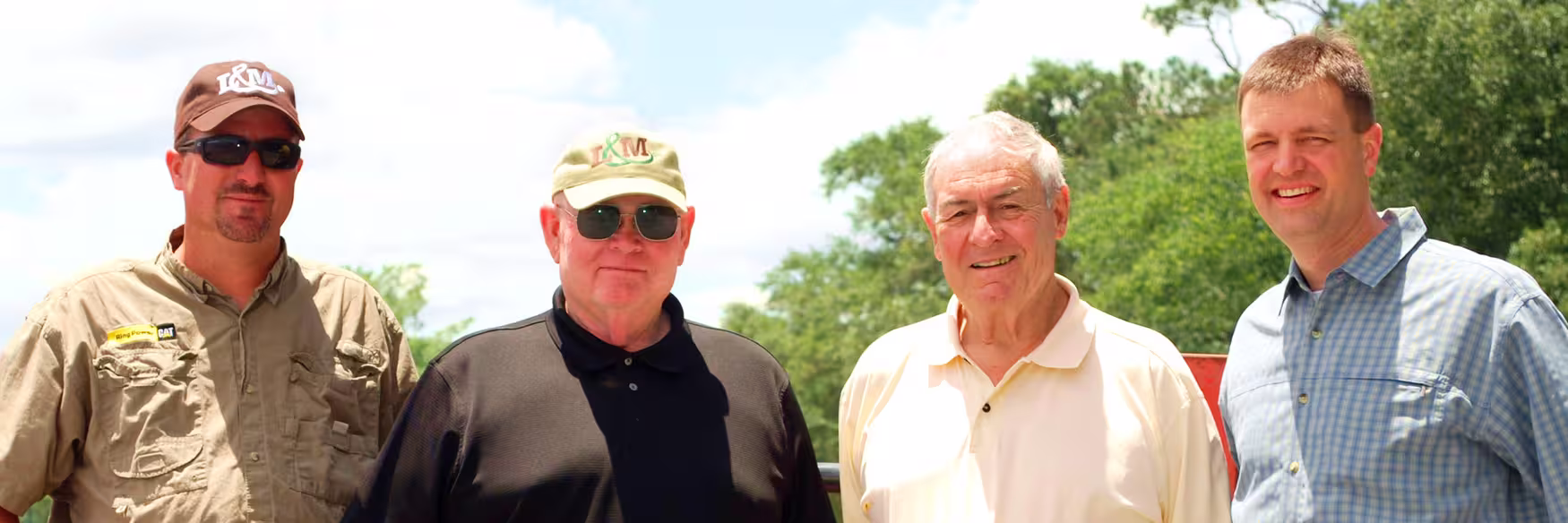Four men standing outdoors in front of green trees and a partly cloudy sky, dressed casually with two wearing sunglasses and hats.