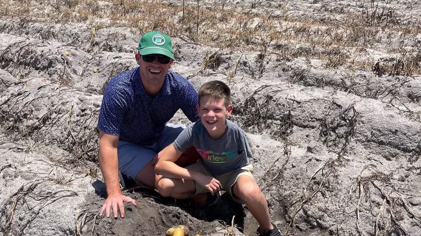 A man wearing a green cap and sunglasses and a boy in a grey t-shirt crouching together in a dry, harvested field with soil mounds and a few potatoes on the ground.