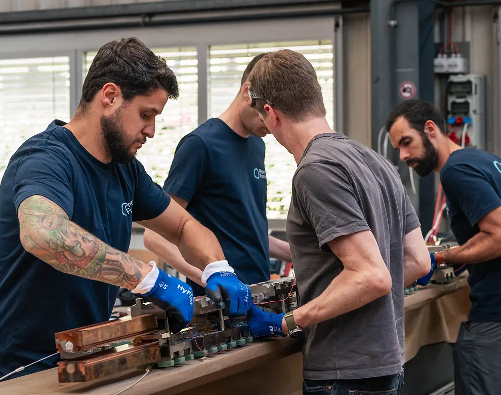Four Proxima Fusion Engineers in gloves working on a piece of hardware while standing.