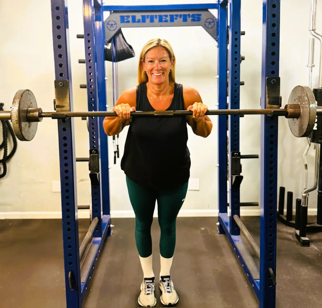 Woman smiling while standing with a barbell in a squat rack at LB Fit gym.