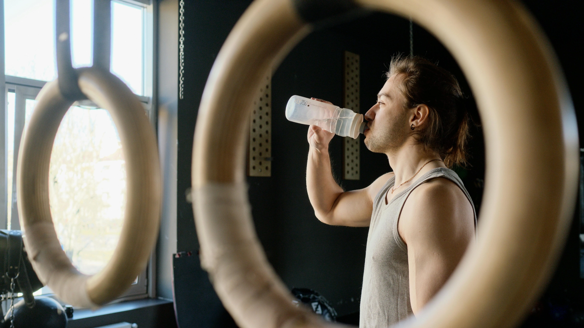 Man drinking water beside gymnastic rings during a workout break at the gym.