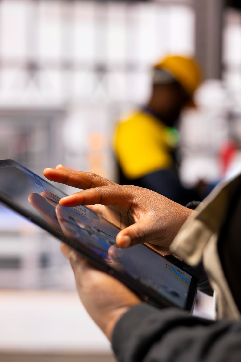 Hands using a tablet on a warehouse floor, with a worker in a hard hat in the background