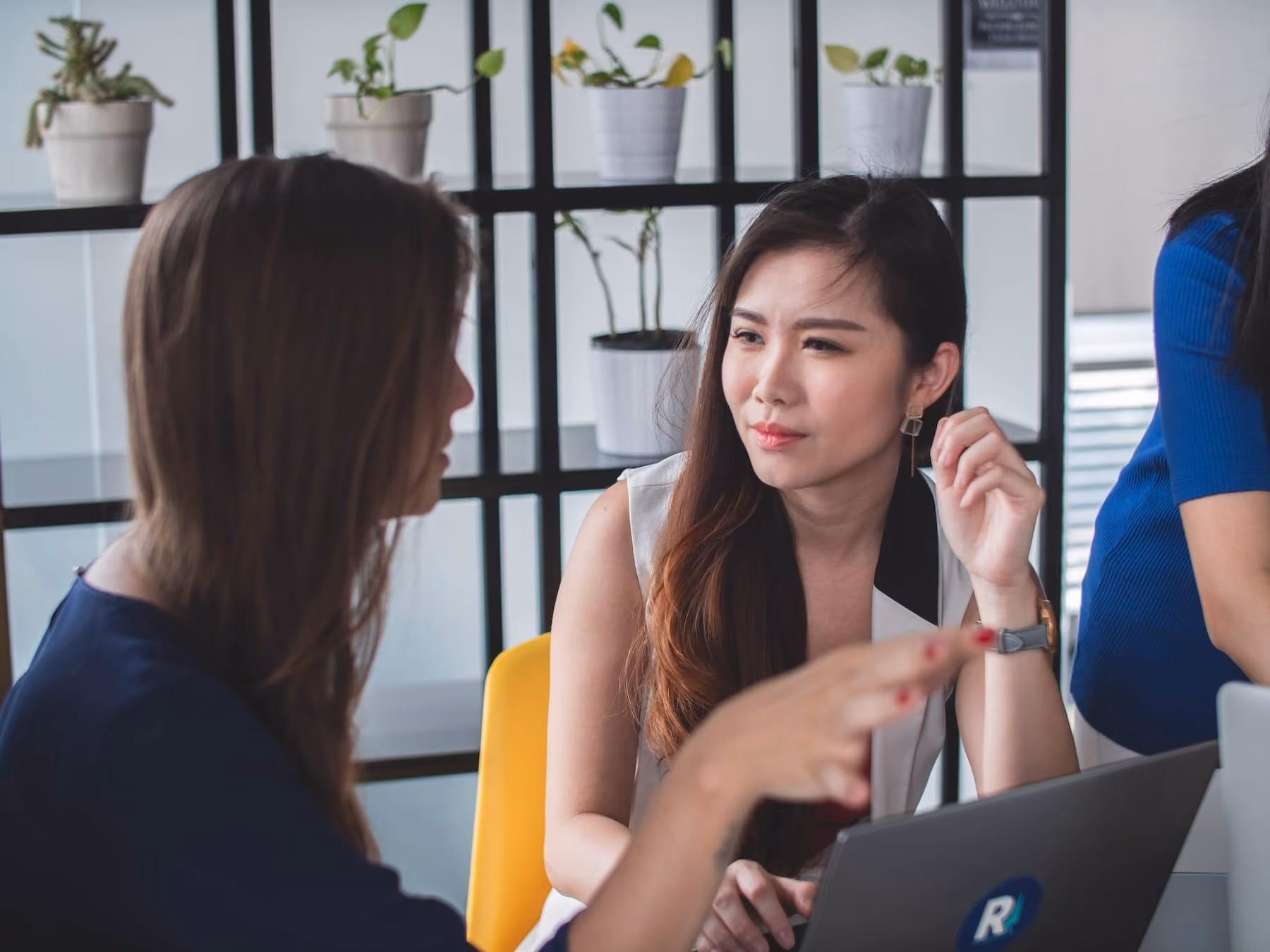 Two women in an office discussing something while looking at a laptop on a table.