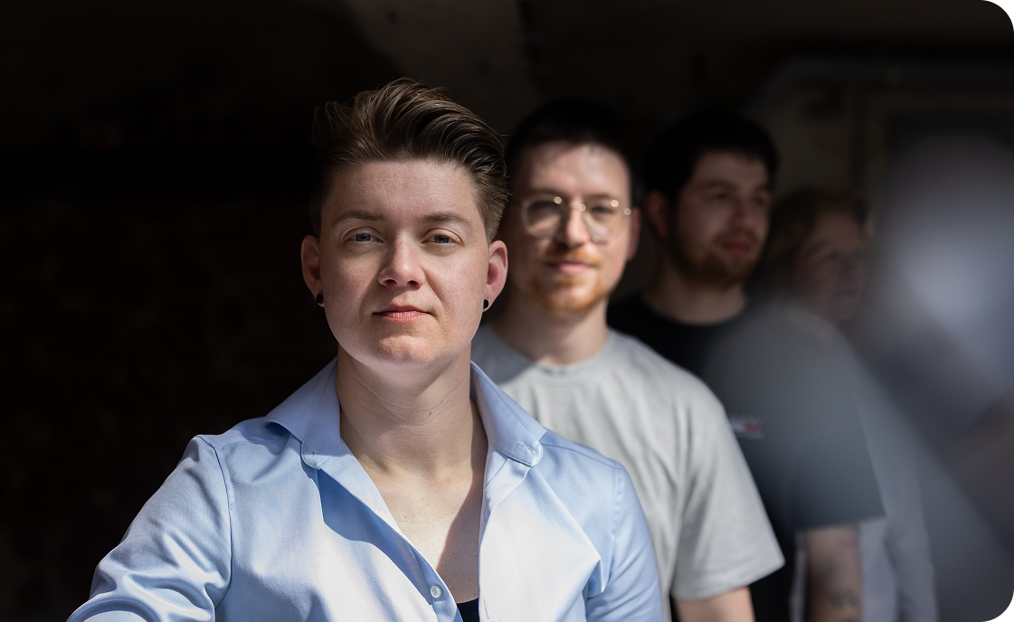 Four people standing in a staggered line, facing forward, against a dark background, with the person at the front wearing a light blue shirt in sharp focus.