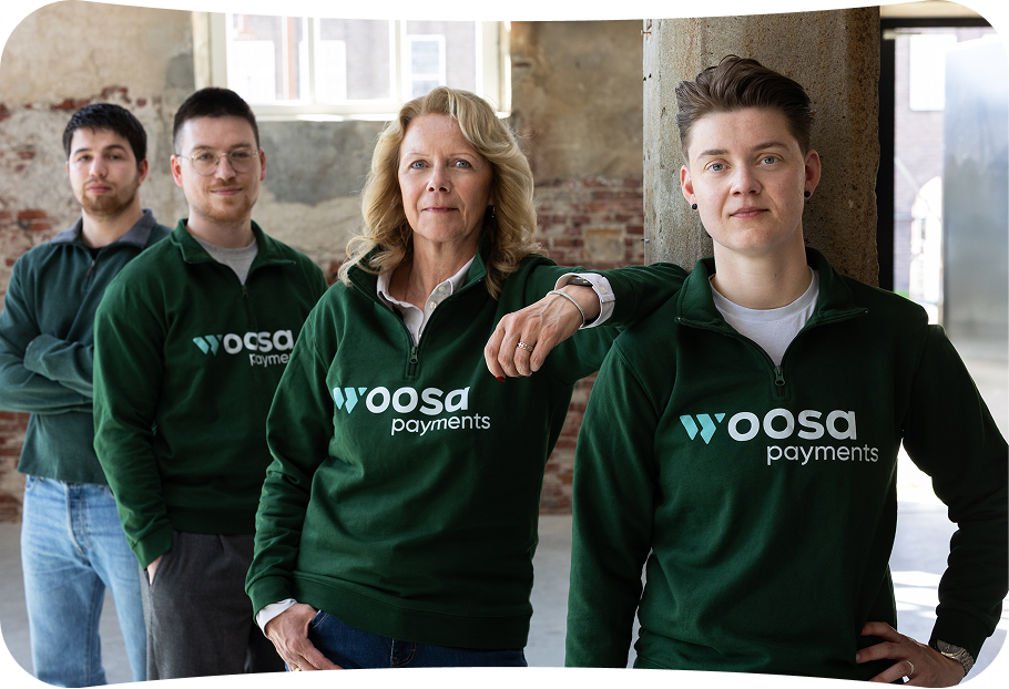 Four people standing in a staggered line indoors, all wearing green Woosa Payments sweatshirts, with a brick wall and windows in the background.