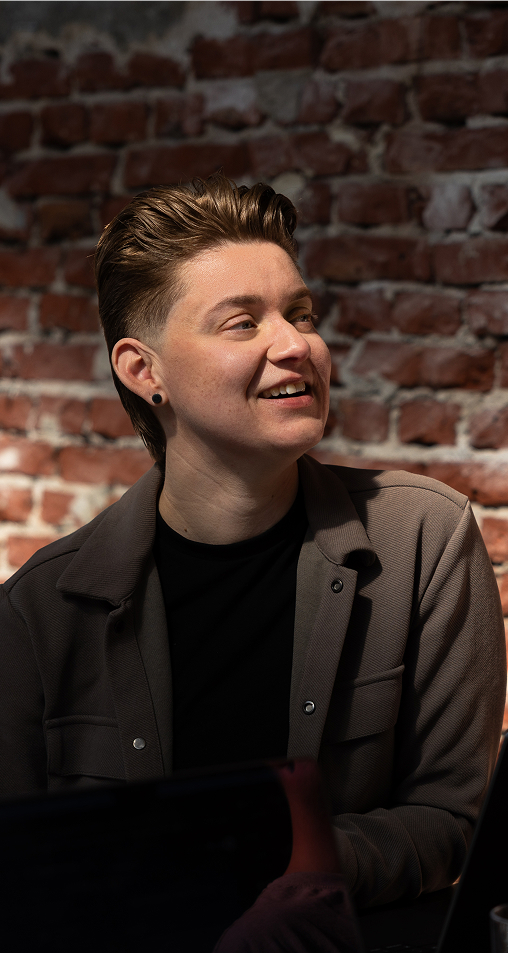 Person with short styled hair, wearing a black shirt and gray jacket, smiling and looking slightly to the left, seated in front of a red brick wall.