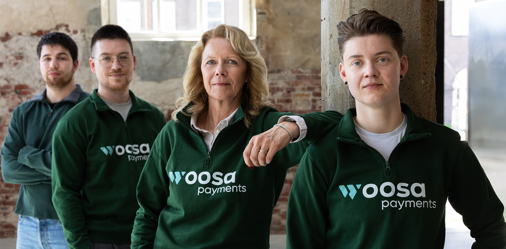 Four people standing indoors against a brick wall, wearing dark green Woosa Payments quarter-zip sweatshirts, looking confidently at the camera.