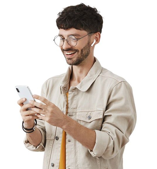 Smiling man with curly dark hair, beard, and round glasses wearing a beige jacket and orange shirt, looking at and holding a smartphone.