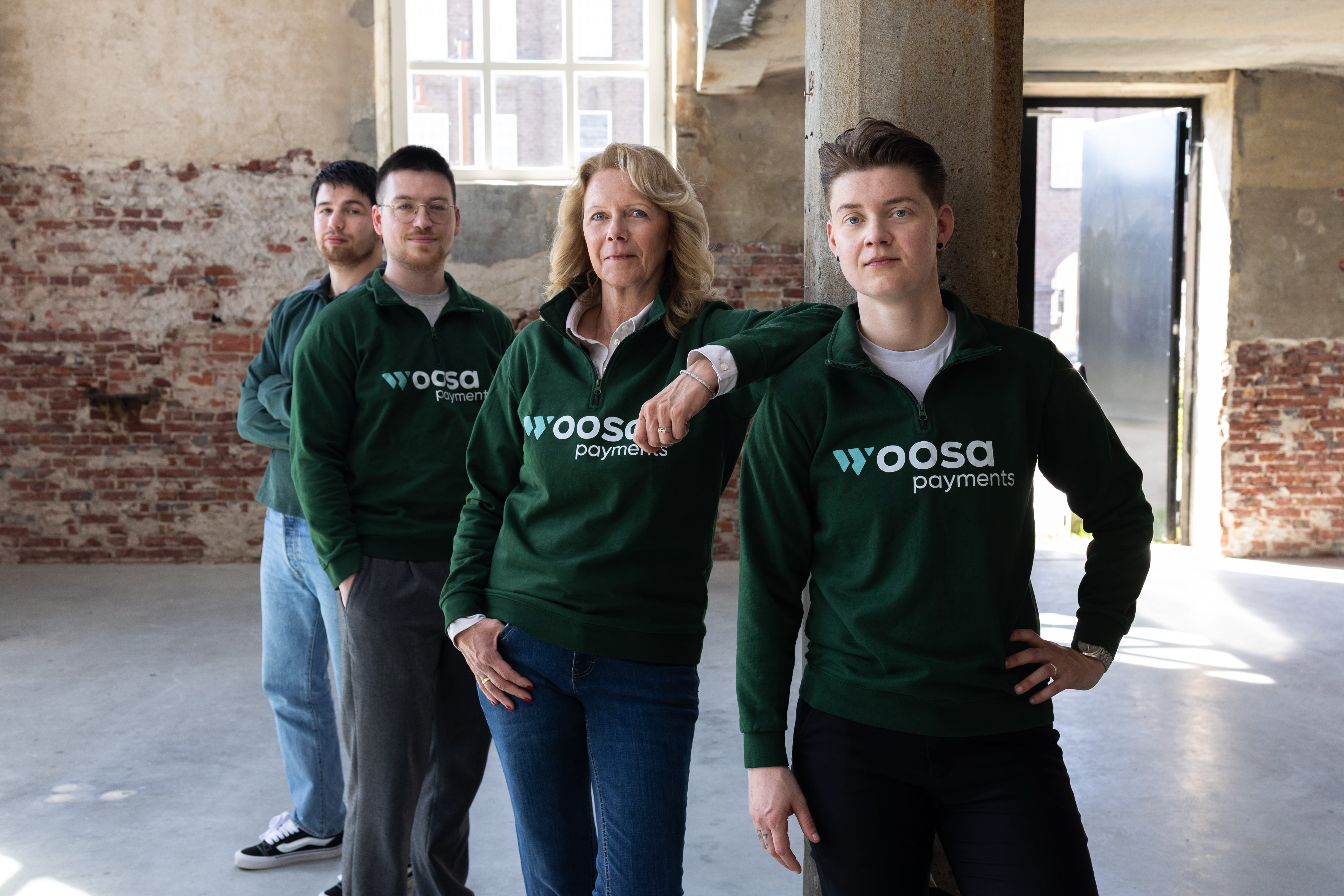 Four people standing in a row indoors in a spacious, unfinished room with brick walls and large windows; three wear dark green sweatshirts with Woosa Payments logo, the woman second from the right rests her arm on the shoulder of the person on the far right.