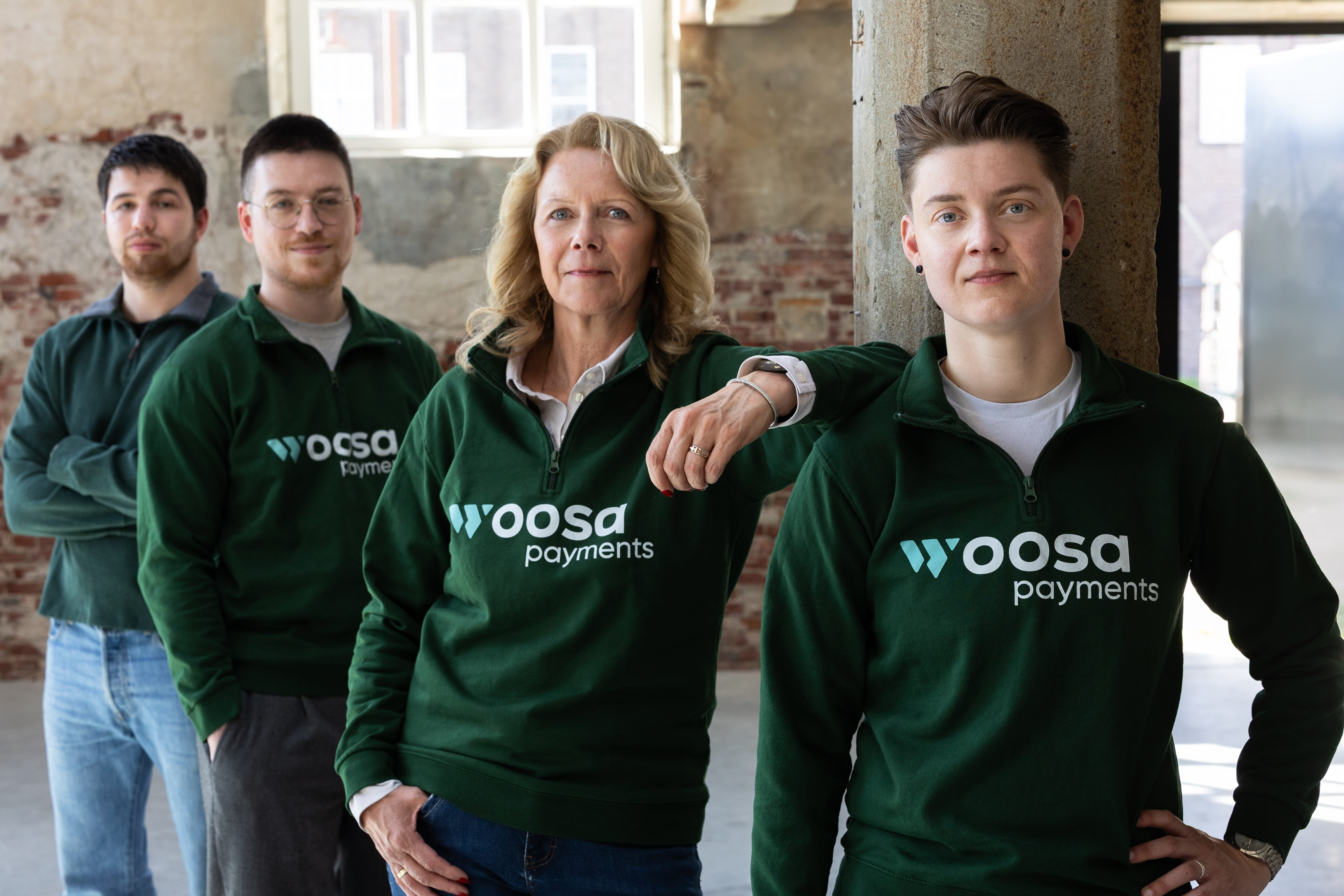 Four people standing indoors against a rustic brick and stone wall, all wearing dark green sweatshirts with the white Woosa Payments logo, looking confidently at the camera in a staggered formation from left to right.