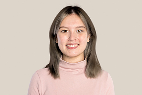 Smiling young woman with shoulder-length straight brown hair wearing a light pink turtleneck top against a plain beige background.