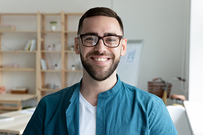 Smiling man with glasses and short dark hair wearing a teal jacket over a white shirt, sitting in a well-lit modern office space with shelves and a desk in the background.