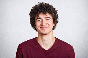 Young man with curly dark hair wearing a maroon V-neck shirt smiling against a plain light gray background.