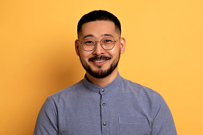 Smiling man with short black hair, beard, and round glasses wearing a buttoned gray shirt against a plain orange background.