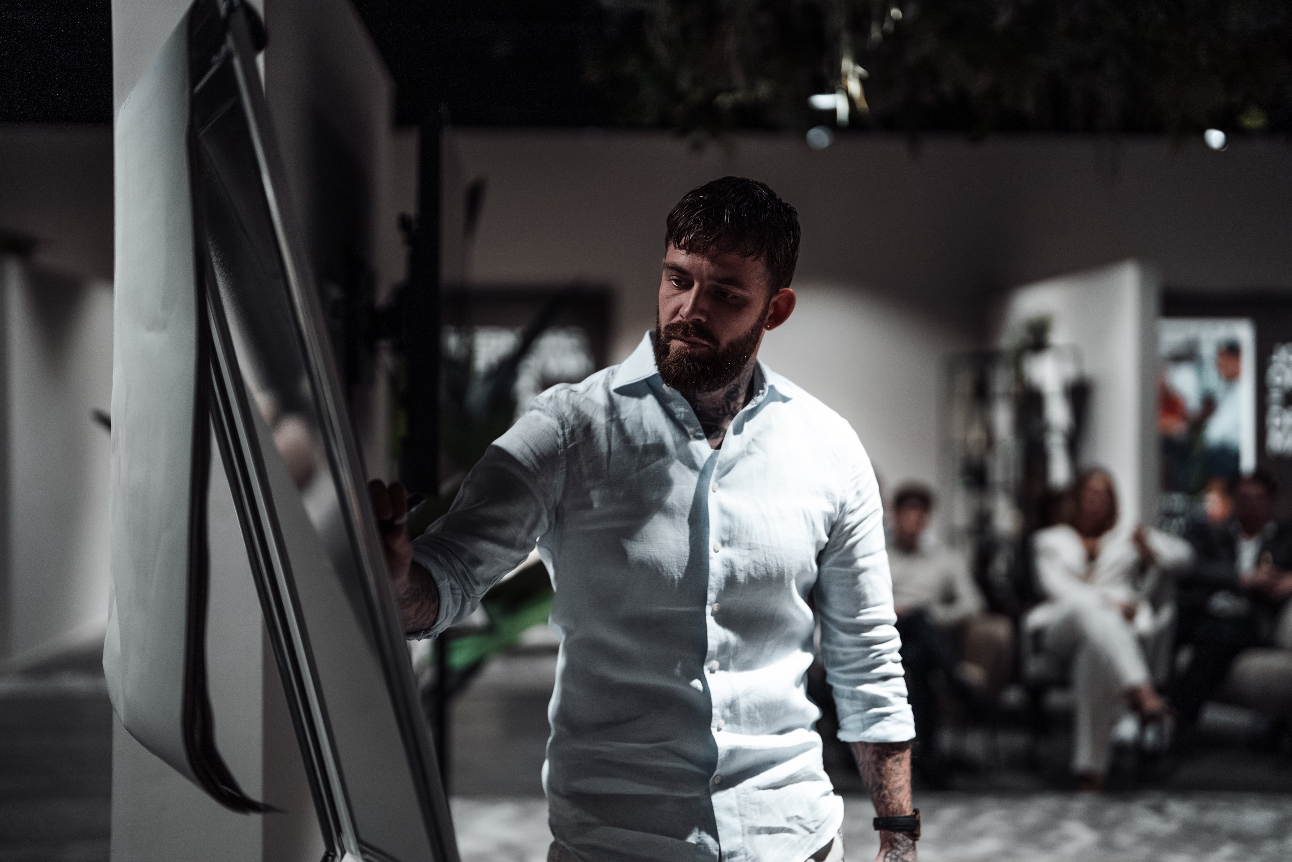 A bearded man wearing a white shirt writes on a flip chart or whiteboard in a dimly lit room while a small group of people seated in the background watch attentively.