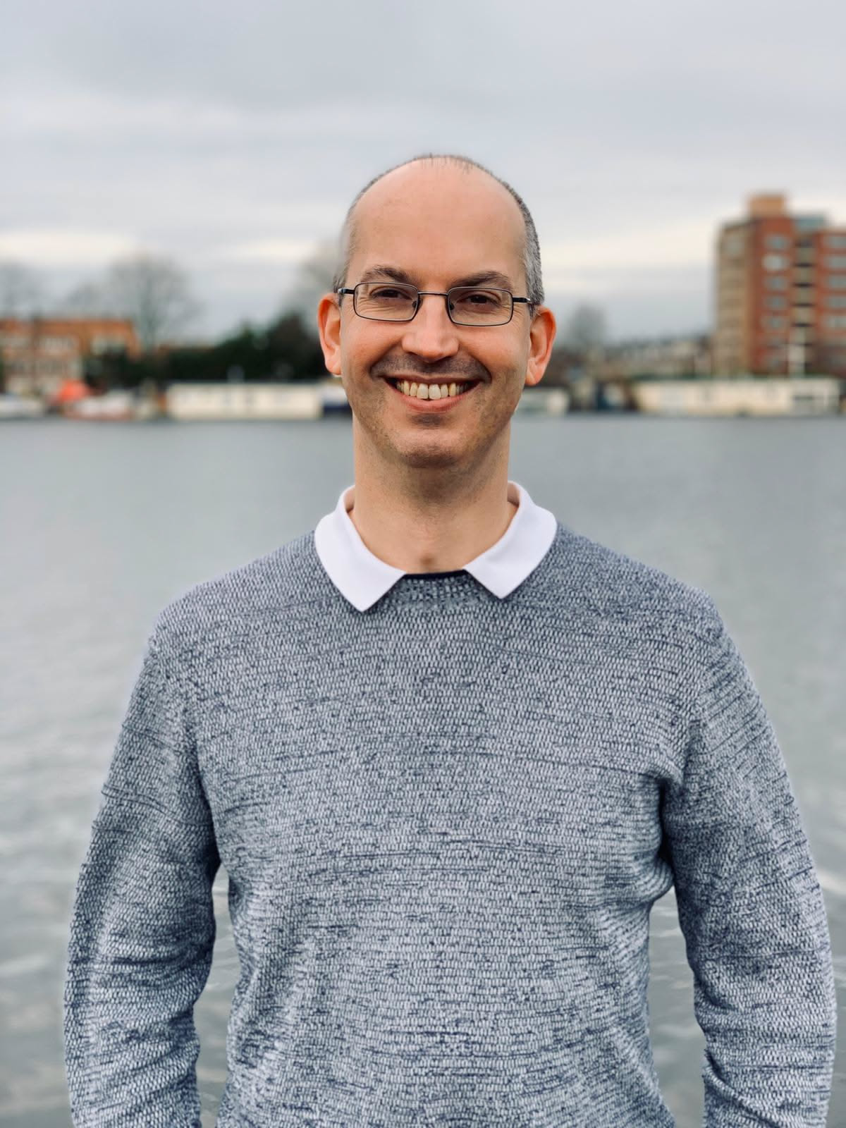 Smiling middle-aged man with glasses wearing a gray sweater over a white collared shirt standing in front of a body of water with buildings and trees in the background under a cloudy sky.