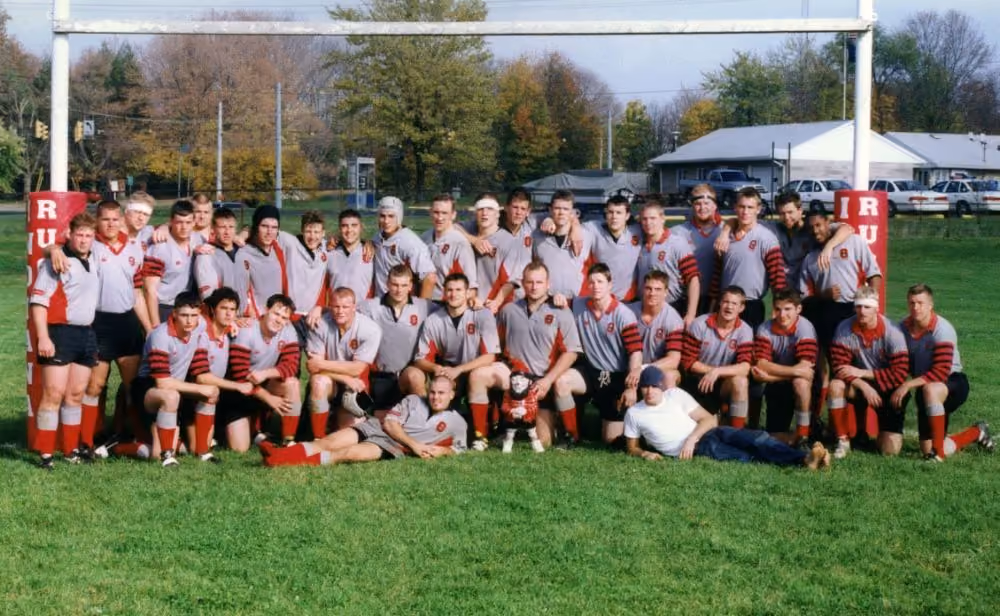 Group photo of a men's rugby team in gray and red uniforms on grass under goalposts.