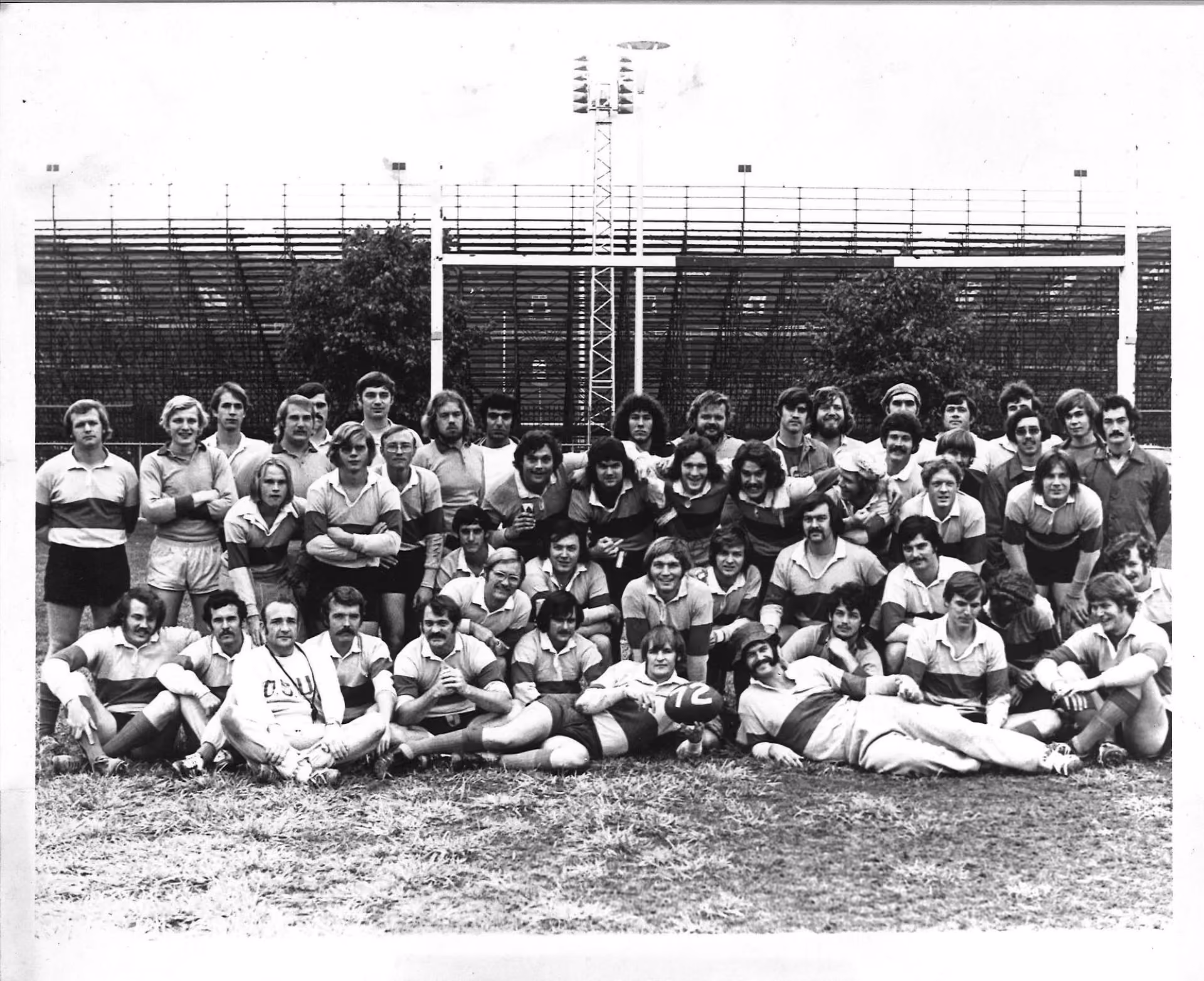 Large group of rugby players and coaches posing on a field in front of goalposts and bleachers.