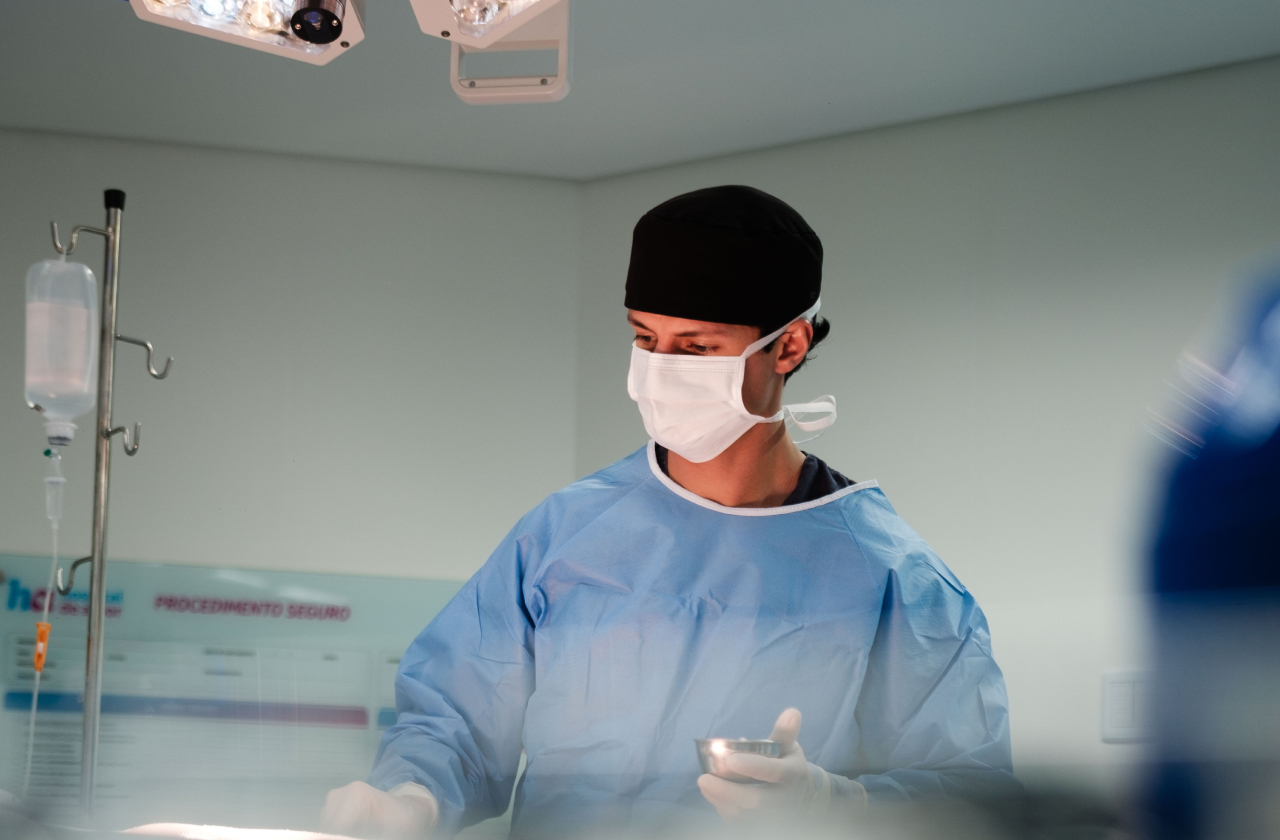 Surgeon in blue scrubs, surgical gloves, black cap, and white face mask holding a small metal container in an operating room under surgery lights.