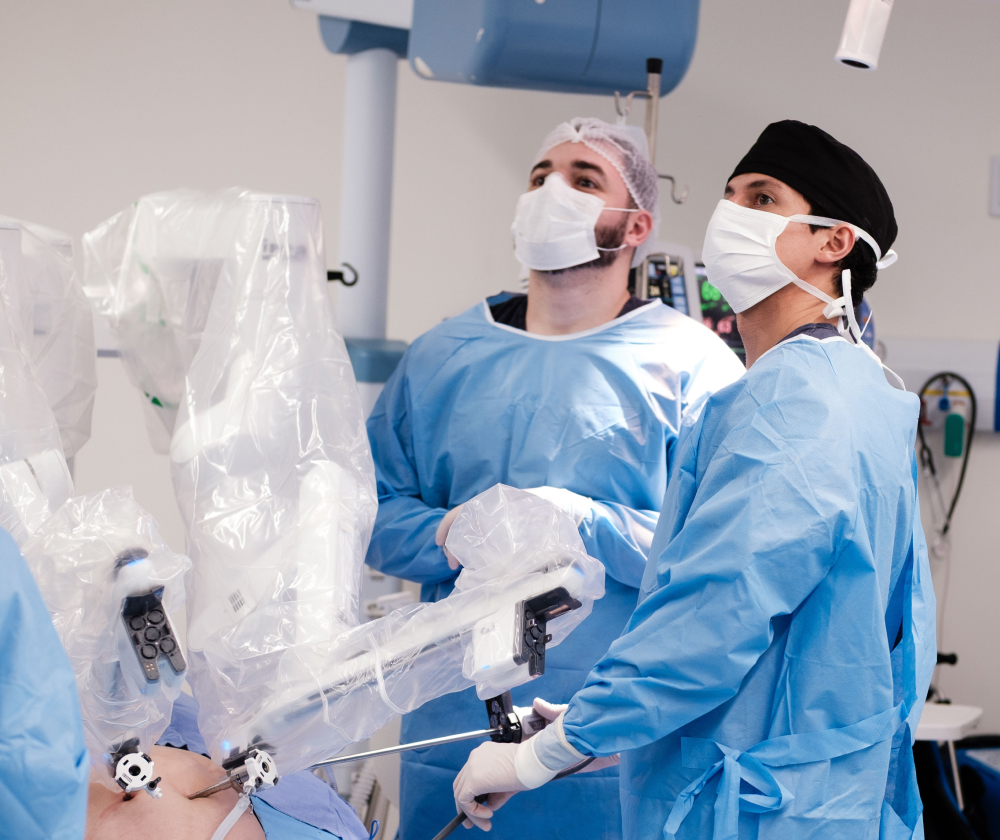 Two surgeons in blue sterile gowns, white face masks, and hair covers operate using a robotic surgical system with multiple arms inside an operating room.
