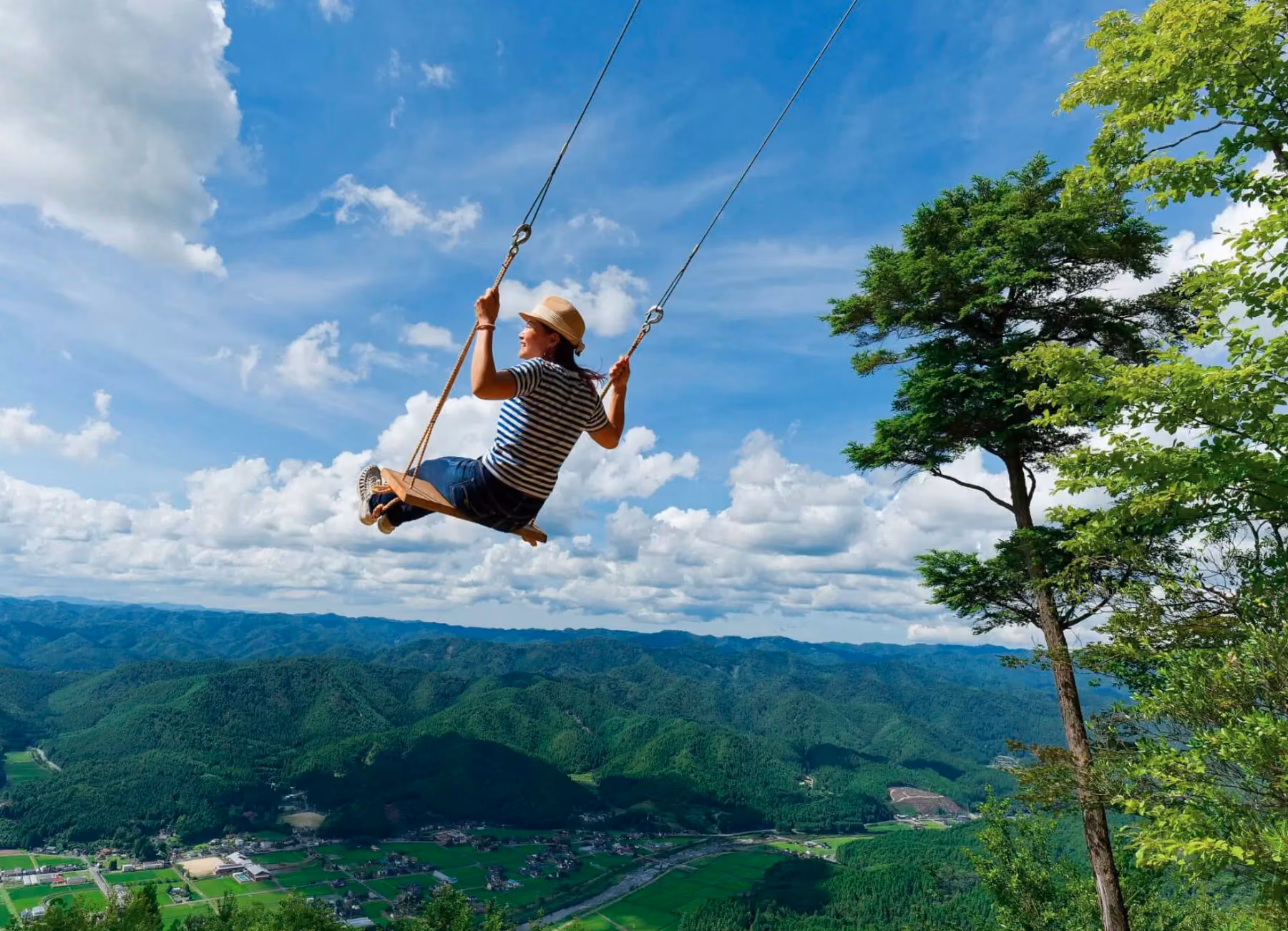 Woman wearing a hat swings high over a lush green valley with blue sky and clouds.