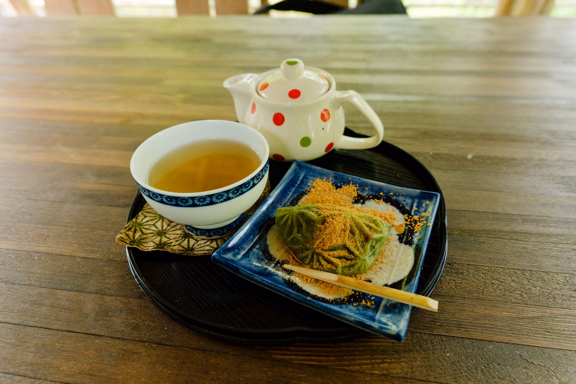 Tray with a teapot, a cup of tea, and a green leaf-shaped sweet sprinkled with brown powder.