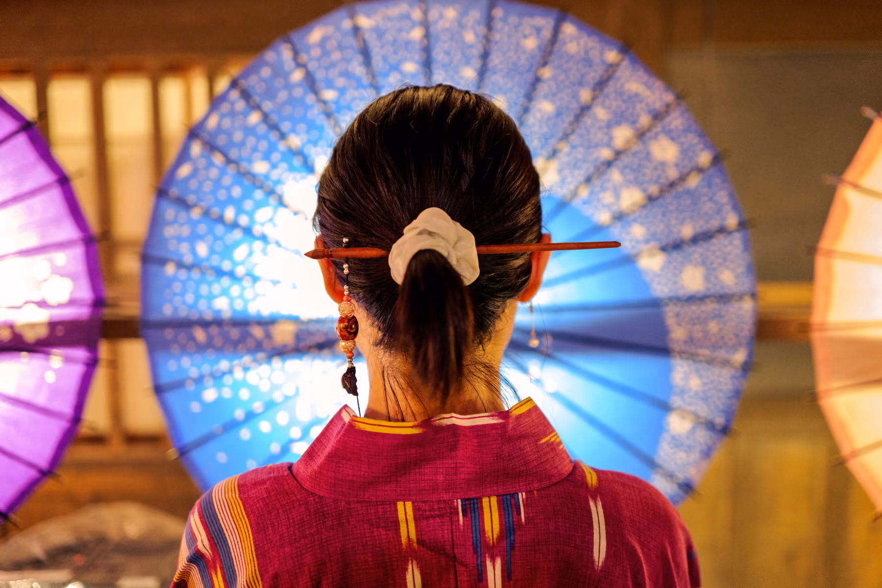 Woman's back with a hair stick and scrunchie, wearing a pink robe, with colorful umbrellas in the background.