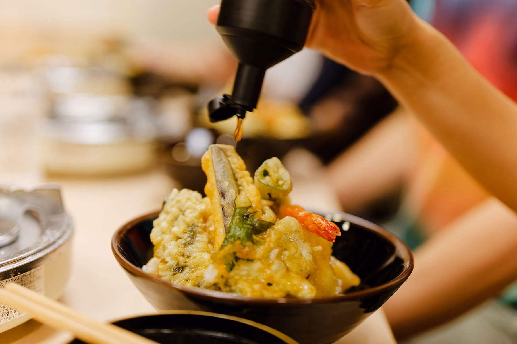 Hand pouring sauce onto assorted tempura vegetables and shrimp in a black bowl.