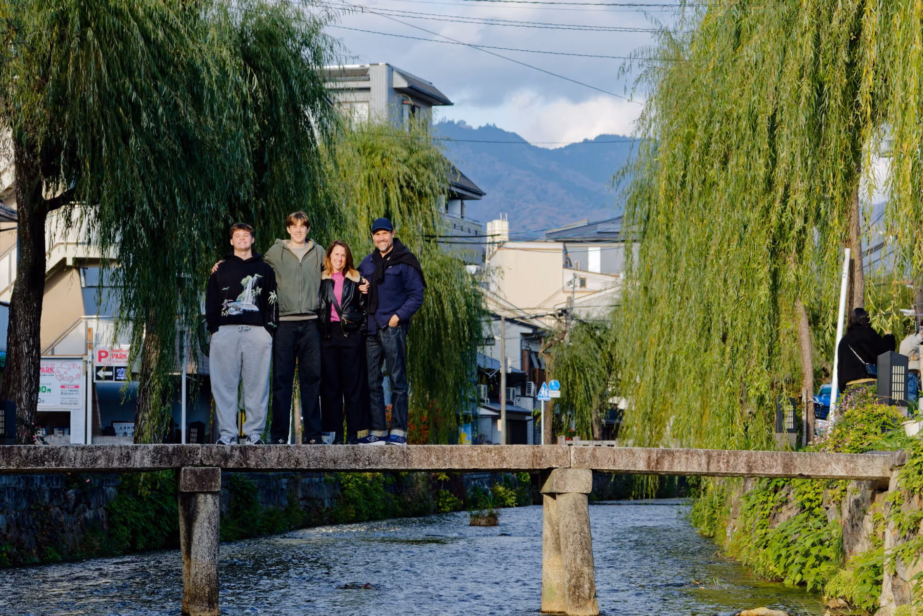 Four people standing closely on a stone bridge over a river, with trees and buildings behind them.