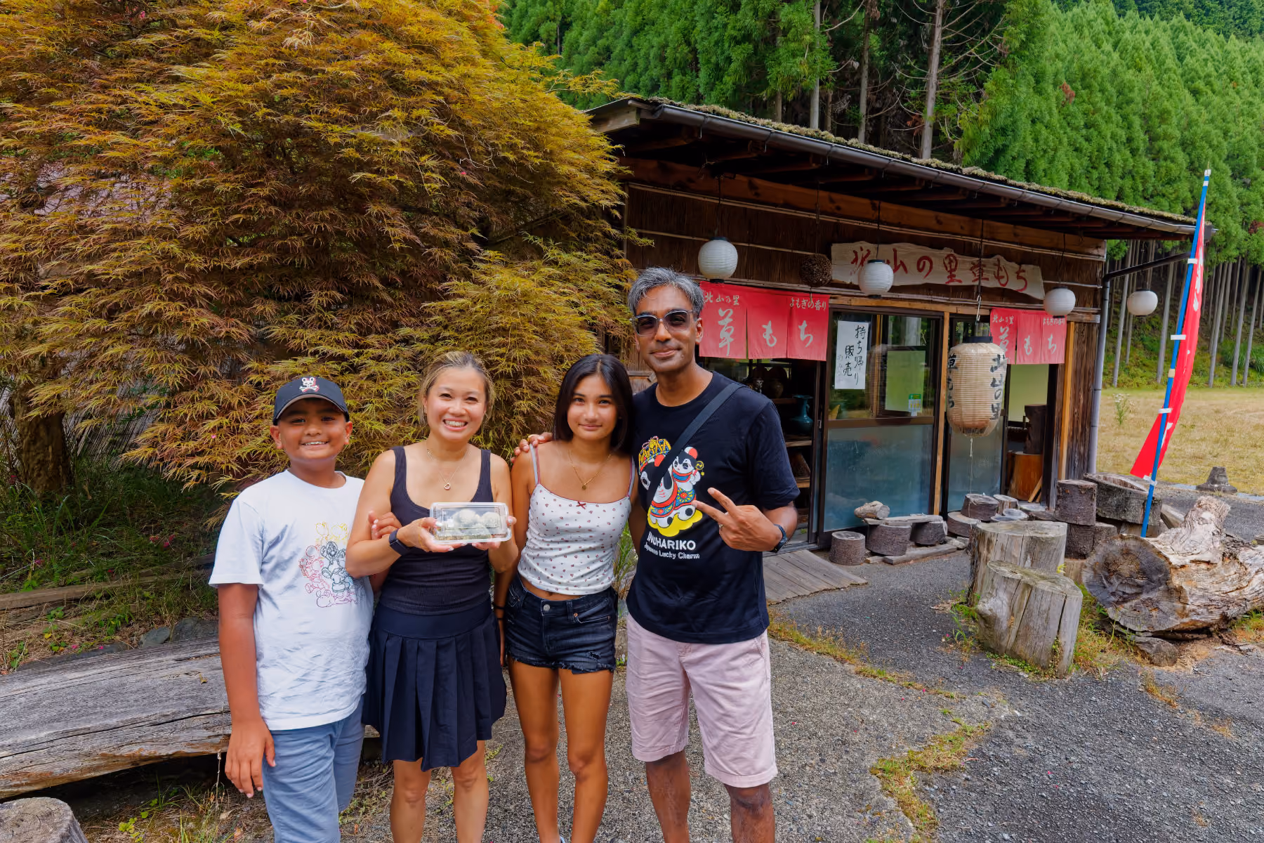 Smiling family of four posing outside a small Japanese shop with wooden decor and greenery around.